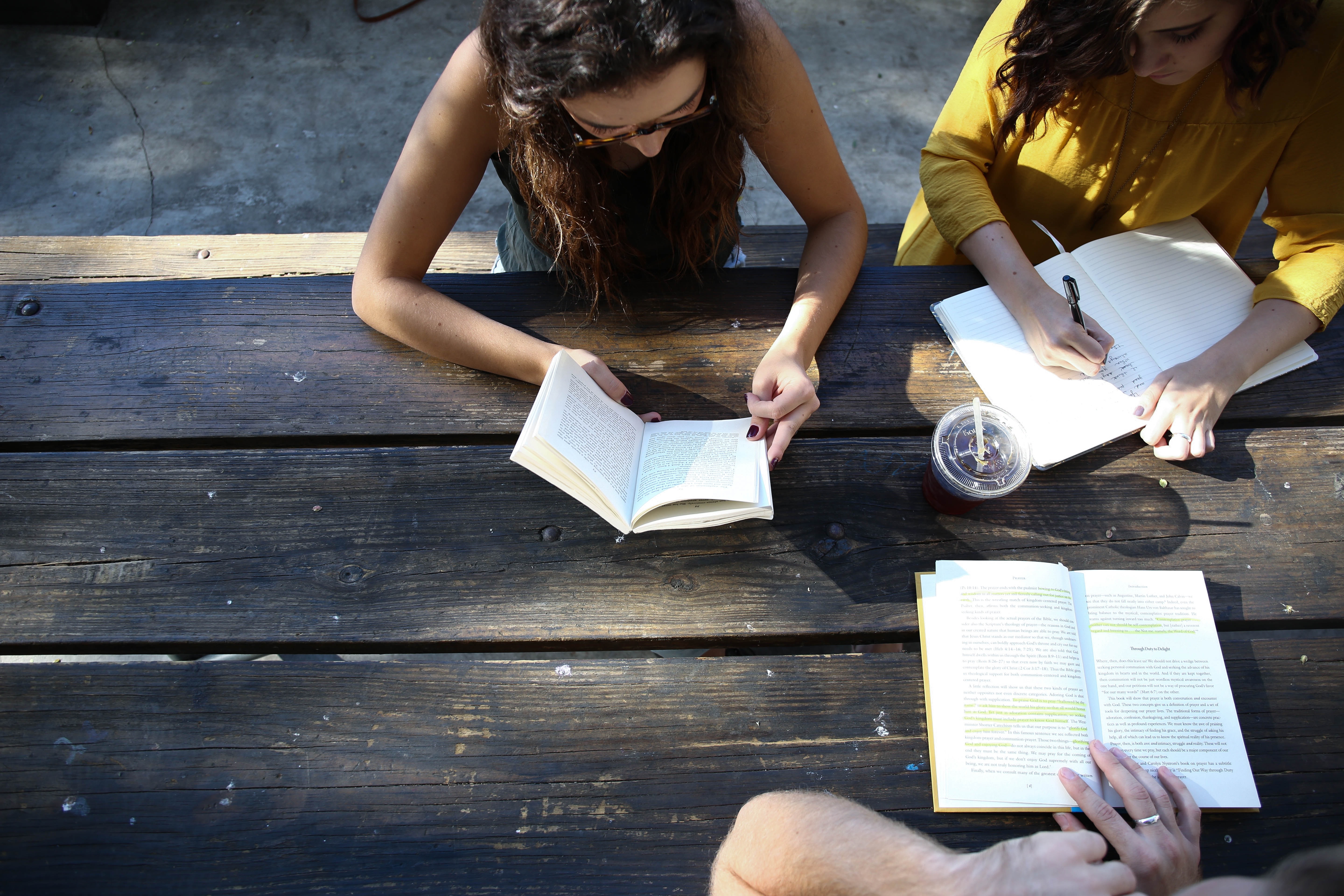Three young women study at a picnic table. Each holds a book, and two are reading. A third takes notes on the pages in front of her. A drink in a clear plastic cup sits to her right.